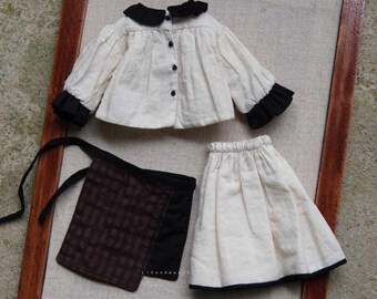 May include: A doll-sized outfit featuring a cream-colored jacket with black collar and cuffs, a matching skirt, a brown apron, and brown boots. The jacket has black buttons. The skirt and jacket have black trim. The boots have a distressed look.