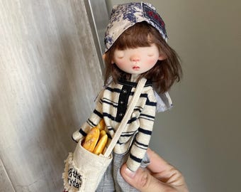 May include: A small doll wearing a striped shirt, a blue and white patterned hat, and gray pants. The doll is holding a bag with the words "of the everyday" embroidered on it. The bag is filled with bread sticks.