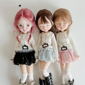May include: Three dolls with closed eyes, each with different hair colours: pink, brown, and blonde. They wear white long-sleeved shirts, skirts in black, blue, and pink, and black boots. Each doll has a tag around their neck.