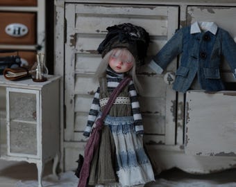 May include: A doll with white hair and a black and white striped shirt, wearing a brown and blue skirt and a brown hat. The doll is standing in front of a white cabinet with a blue jacket hanging on it.
