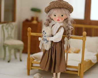 Könnte beinhalten: Eine Puppe mit blonden Zöpfen, die einen braunen Rock, eine weiße Bluse und einen braunen Strohhut trägt, hält ein weißes Stofftier. Die Puppe steht auf einem Holzbettgestell mit einer weißen Decke.