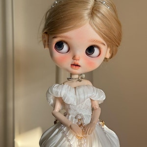 Op de afbeelding: Een Blythe-pop met blond haar en grote, expressieve ogen. De pop draagt een witte, gelaagde jurk met een parelketting en een met parels versierde haarband. Een streng parels wordt in de handen van de pop gehouden, wat bijdraagt aan de elegante uitstraling.