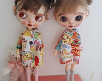 Puede incluir: Dos muñecas con ojos grandes y cabello castaño peinado en moños. Llevan vestidos coloridos con ilustraciones de dibujos animados, calcetines rosas y zapatos amarillos. Una muñeca tiene un osito de peluche al lado.