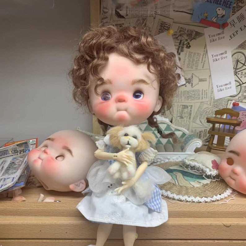 May include: A doll with curly brown hair wearing a blue and white dress and holding a small white stuffed dog. The doll has blue eyes and rosy cheeks.
