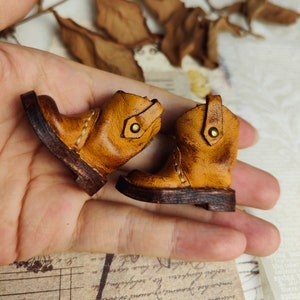 May include: A pair of miniature brown leather boots with a distressed finish. The boots have a pointed toe, a buckle on the side, and a brown leather sole.