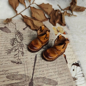 May include: A pair of miniature brown leather boots with a pointed toe and a stitched seam around the sole. The boots are on a patterned background.