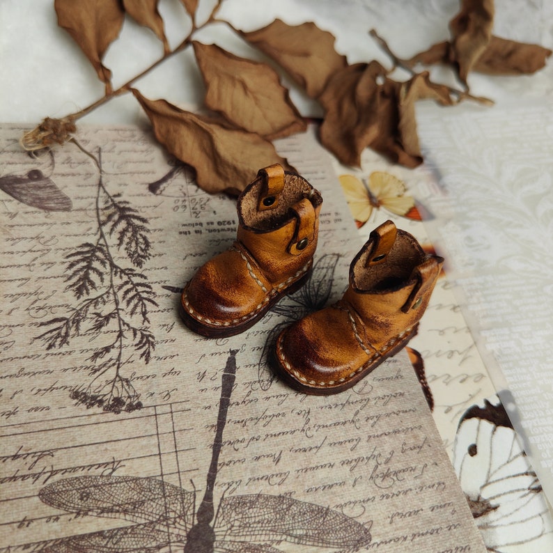May include: A pair of miniature brown leather boots with a pointed toe and a stitched seam around the sole. The boots are on a patterned background.