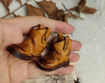 May include: A pair of miniature brown leather boots with stitching detail and a button accent. The boots are on a hand.