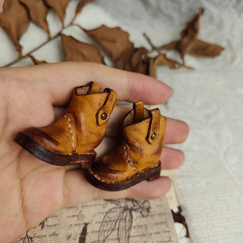 May include: A pair of miniature brown leather boots with stitching detail and a button accent. The boots are on a hand.