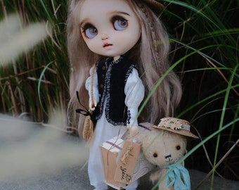 May include: A doll with long blonde hair wearing a black vest and a white dress, holding a brown box with the text "I love you" on it. The doll is standing next to a small, furry, brown teddy bear wearing a straw hat with flowers.