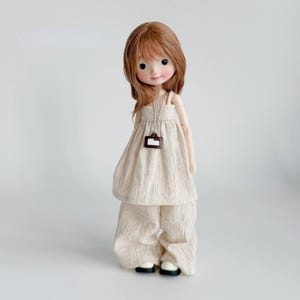May include: A doll with light brown hair and rosy cheeks wearing a cream-coloured linen outfit. The outfit includes a top with a square pendant necklace and wide-leg trousers. The doll is standing and wearing black and white shoes.