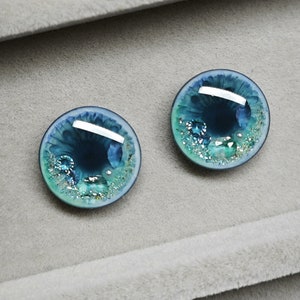 May include: Two round blue and green glass eyes with glitter and silver accents. The eyes are set against a gray background.
