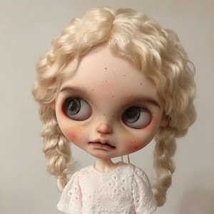 Może przedstawiać: Lalka porcelanowa z blond, kręconymi włosami ułożonymi w dwa warkocze. Lalka ma niebieskie oczy i jest ubrana w białą sukienkę z koronki.