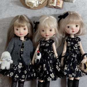 May include: Three dolls with blonde and light brown hair are dressed in black floral dresses and accessories. One doll wears a grey cardigan and holds a white stuffed animal. Another doll holds a small bird. The third doll carries a small handbag.