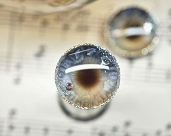 May include: Close-up of a pair of handmade glass eyes with silver glitter accents. The eyes are brown and gray with a white iris and a black pupil. The eyes are set on a white background.