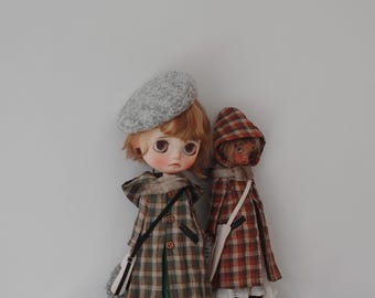 Könnte beinhalten: Zwei Puppen in Kleidung im Vintage-Stil stehen nebeneinander. Die Puppe links trägt eine graue Baskenmütze, einen karierten Mantel mit grünen Akzenten und eine Umhängetasche. Die Puppe rechts trägt einen karierten Kapuzenmantel. Beide Puppen haben detaillierte Gesichtszüge.