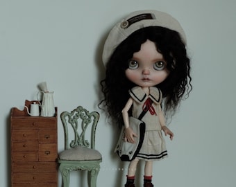 May include: A doll with long black curly hair wearing a white sailor dress with a red bow and a white beret with a brown patch. The doll is holding a white bag with a black strap. The doll is standing on a wooden floor in front of a wooden dresser with drawers.