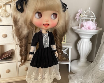 Puede incluir: Una muñeca con un vestido negro con una capa de encaje blanco y lazos negros en el pelo. La muñeca tiene el pelo rubio y los ojos azules.