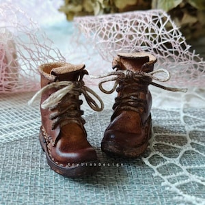 Puede incluir: Un par de botas de cuero marrón en miniatura con cordones. Las botas tienen un aspecto desgastado y vintage, con costuras visibles y una superficie texturizada. Los cordones están atados con un lazo. Las botas son probablemente para una muñeca u otra figura pequeña.