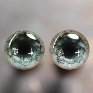 Op de afbeelding: Een paar groene en witte poppenogen met zilveren glitter en een heldere strass-accent. De ogen hebben een zwarte pupil en een witte iris.