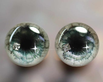 May include: A pair of green and white doll eyes with silver glitter and a clear rhinestone accent. The eyes have a black pupil and a white iris.