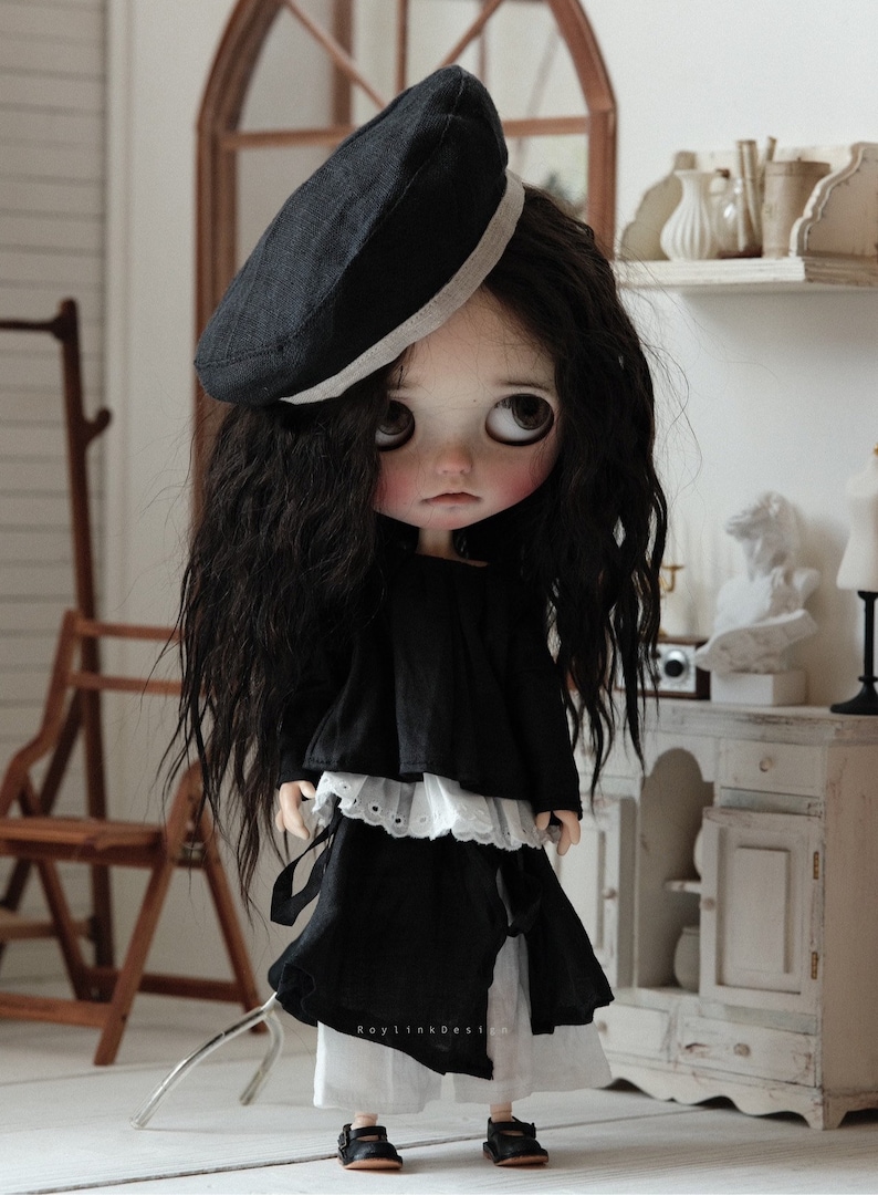 Pu&ograve; includere: Una bambola con lunghi capelli neri che indossa un berretto nero con un bordo bianco, un vestito bianco e nero e pantaloni bianchi. La bambola &egrave; in piedi su una superficie bianca.