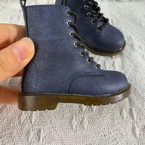 May include: A pair of navy blue ankle boots with a subtle glitter finish, featuring black laces, silver-toned metal eyelets, a side zipper, and a brown rubber sole with a treaded bottom. The boots have a structured design with a pull tab at the heel.