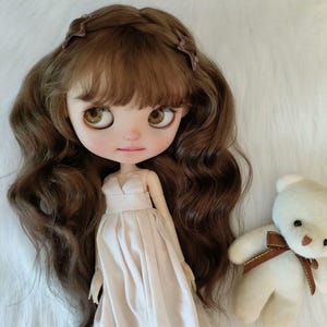 Puede incluir: Una muñeca con cabello castaño y un vestido rosa pálido. La muñeca tiene ojos grandes y una expresión suave. Un pequeño oso de peluche blanco con una cinta marrón está cerca. El cabello de la muñeca está peinado con una trenza.