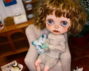 May include: A doll with curly brown hair sits in a light gray armchair holding a small teal teacup. The doll is wearing a light beige sweater and matching shorts. A small white cat sits on the floor next to the chair.