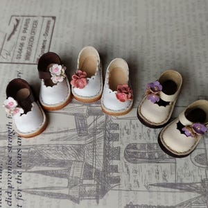 May include: Six pairs of miniature doll shoes. The shoes are white with brown accents and adorned with small, colorful floral embellishments. The shoes are arranged on a patterned surface.