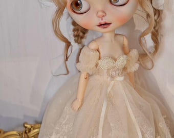 May include: A porcelain doll with blonde hair wearing a white lace dress and a white lace headpiece. The doll has blue eyes and a painted smile.