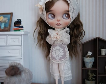 May include: A porcelain doll with long brown hair wearing a white lace dress and a white lace bonnet. The doll is standing on a white lace doily with a small stuffed sheep in the foreground.