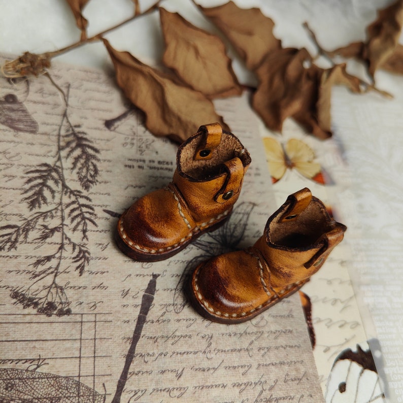 May include: A pair of miniature brown leather boots with white stitching. The boots are sitting on a piece of paper with writing on it.