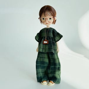 K&ouml;nnte beinhalten: Eine Puppe in einem gr&uuml;n-schwarz karierten Outfit, Brille und einem kleinen braunen Anh&auml;nger. Die Puppe hat braune Haare und tr&auml;gt ein passendes kariertes Oberteil und eine Hose. Die Puppe tr&auml;gt auch kleine, hellbraune Schuhe.