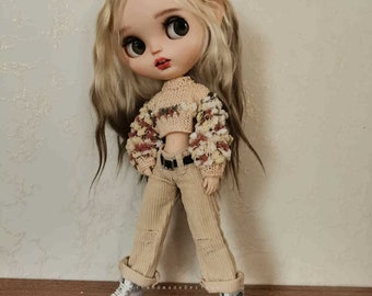 May include: A doll with blonde hair wearing a beige sweater with floral accents, beige corduroy pants, and white sneakers.