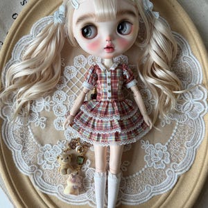 Op de afbeelding: Een Blythe-pop met lang blond haar in vlechtjes, gekleed in een geruite jurk met een gerimpelde rok, witte sokken en zwarte schoenen. De pop heeft grote ogen en roze wangen. Een bedel met een teddybeer en andere kleine figuurtjes hangt aan de pop.