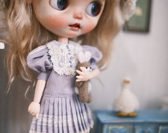 May include: A doll wearing a lavender dress with a white lace collar and a white lace trim. The doll is holding a small stuffed bunny in its right hand. The doll has blonde hair and blue eyes.