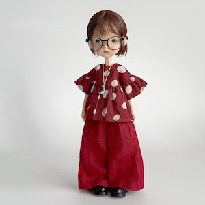 Può includere: Una bambola indossa un top rosso a pois e pantaloni a gamba larga abbinati. La bambola ha i capelli castani corti, occhiali rotondi e una collana con pendente argentato. Le scarpe nere completano l'outfit.