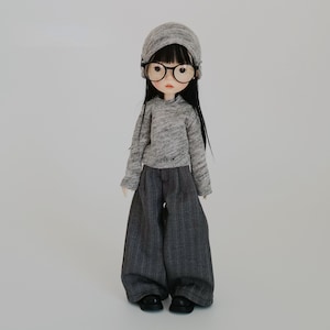 May include: A doll wearing a gray knit beanie, round black glasses, and a matching gray sweater. The doll is also wearing wide-leg gray pants and black shoes. The doll has long black hair and a pale complexion.