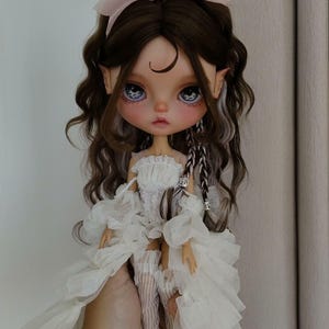 May include: A doll with long brown hair, wearing a white dress with lace detailing and a pink bow in her hair. The doll has blue eyes and a crescent moon painted on her forehead.