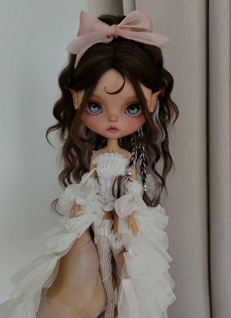 May include: A doll with long brown hair, wearing a white dress with lace detailing and a pink bow in her hair. The doll has blue eyes and a crescent moon painted on her forehead.