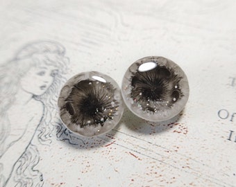 May include: Two round, clear resin earrings with a dark gray, feathery design in the center. The earrings have a speckled, glittery border and are set against a white background with a faint illustration.
