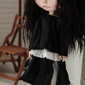 Pu&ograve; includere: Una bambola in bianco e nero con lunghi capelli neri che indossa un vestito nero con un bordo di pizzo bianco e scarpe nere.
