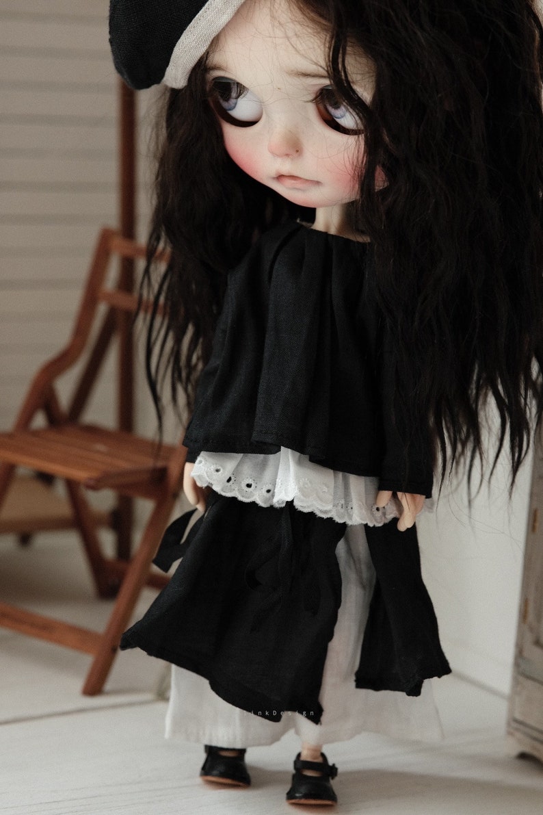 Pu&ograve; includere: Una bambola in bianco e nero con lunghi capelli neri che indossa un vestito nero con un bordo di pizzo bianco e scarpe nere.