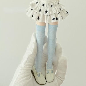 Può includere: Una bambola con una gonna bianca a stelle nere, calze al ginocchio azzurre e scarpe Mary Jane bianche. La gonna ha un design a strati con stelle. Le scarpe hanno un cinturino e un piccolo tacco. Le gambe della bambola sono visibili.