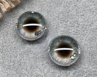 May include: Two round, gray and brown glass eyes with white pupils and silver glitter. The eyes are set against a gray background.