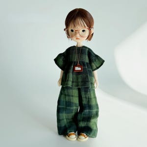 K&ouml;nnte beinhalten: Eine Puppe in einem gr&uuml;n-schwarz karierten Outfit, bestehend aus einem Oberteil, einer weiten Hose und kleinen braunen Schuhen. Die Puppe hat braune Haare, eine Brille und eine Halskette mit einem kleinen Anh&auml;nger. Das Outfit ist aus einem strukturierten Stoff.