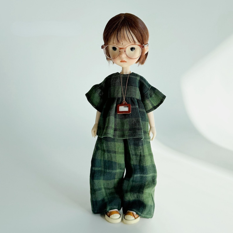 K&ouml;nnte beinhalten: Eine Puppe in einem gr&uuml;n-schwarz karierten Outfit, bestehend aus einem Oberteil, einer weiten Hose und kleinen braunen Schuhen. Die Puppe hat braune Haare, eine Brille und eine Halskette mit einem kleinen Anh&auml;nger. Das Outfit ist aus einem strukturierten Stoff.