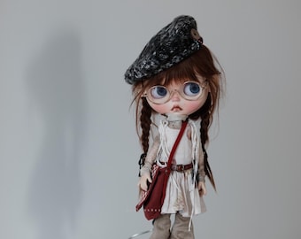May include: A doll with brown hair wearing a black and white beret, round glasses, a white dress with a brown belt, and brown pants. The doll is holding a red bag.