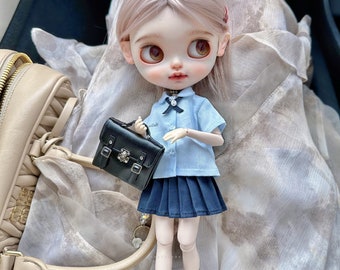 May include: A doll dressed in a light blue school uniform with a pleated navy skirt and black socks. The doll is holding a small black satchel with silver buckles. The doll has light brown eyes and light pink hair.